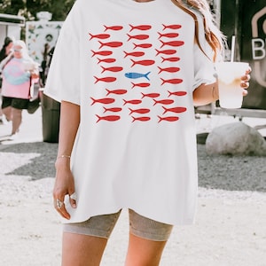 May include: White oversized t-shirt with a graphic of red fish with one blue fish in the middle. The text "WHITE" is at the bottom of the image.