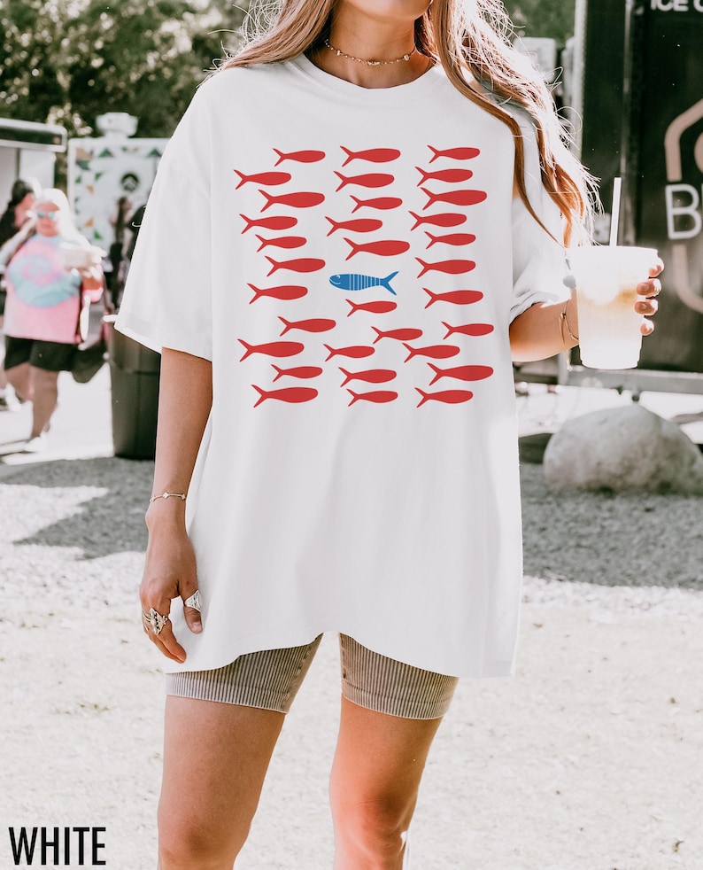 May include: White oversized t-shirt with a graphic of red fish with one blue fish in the middle. The text "WHITE" is at the bottom of the image.