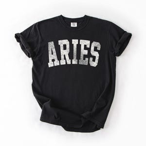 May include: Black t-shirt with the word "ARIES" printed in distressed white lettering. The shirt has short sleeves and a crew neck. The fabric appears soft and comfortable, suitable for casual wear.