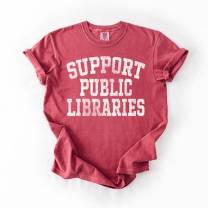 May include: A red t-shirt with rolled-up sleeves. The shirt features the text "SUPPORT PUBLIC LIBRARIES" in a white, distressed font. The shirt is made of a soft material and is likely a unisex fit.