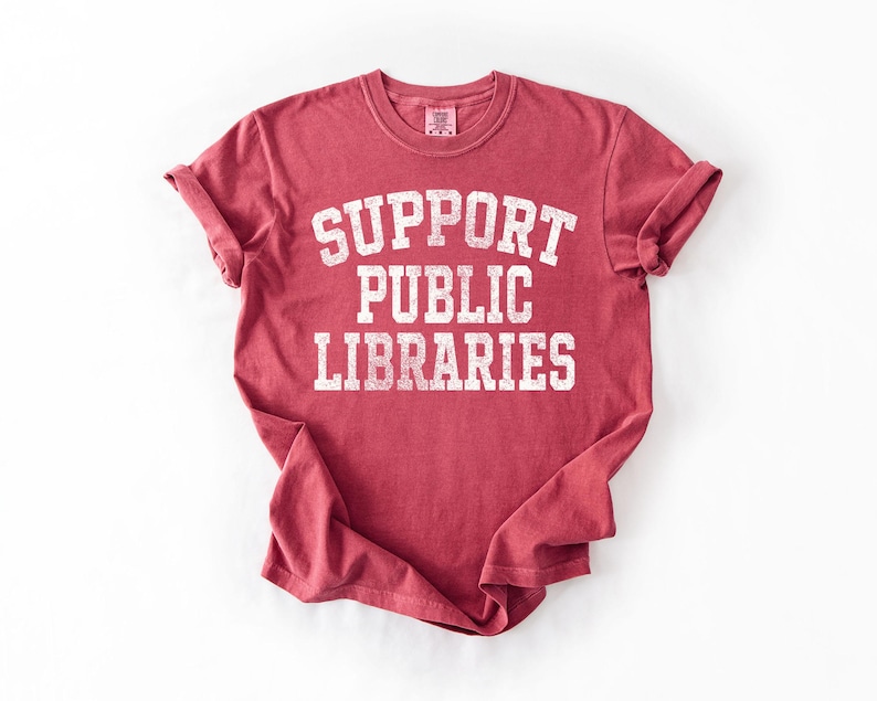 May include: A red t-shirt with rolled-up sleeves. The shirt features the text "SUPPORT PUBLIC LIBRARIES" in a white, distressed font. The shirt is made of a soft material and is likely a unisex fit.