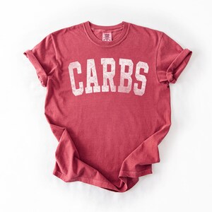 May include: A red t-shirt with the word "CARBS" printed in large, white, distressed letters across the front. The shirt has rolled-up sleeves and a crew neck. The fabric appears soft and worn.
