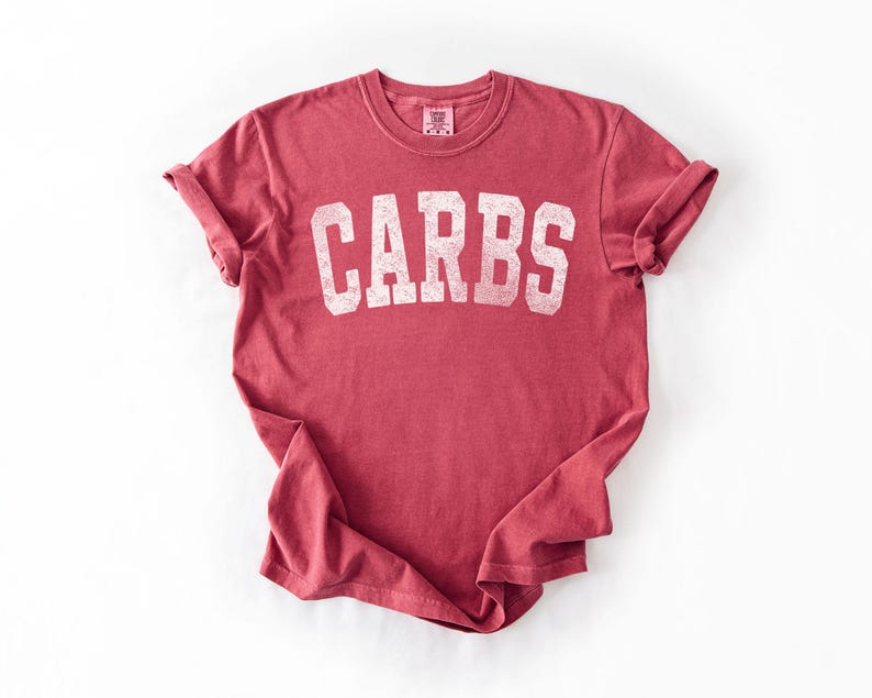 May include: A red t-shirt with the word "CARBS" printed in large, white, distressed letters across the front. The shirt has rolled-up sleeves and a crew neck. The fabric appears soft and worn.