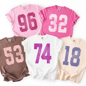 May include: Five short-sleeved t-shirts in various colors: pink, brown, white, and peach. Each shirt features a large, distressed number in a contrasting color. The numbers are 96, 32, 53, 74, and 18.