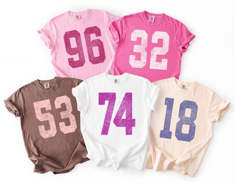 May include: Five short-sleeved t-shirts in various colors: pink, brown, white, and peach. Each shirt features a large, distressed number in a contrasting color. The numbers are 96, 32, 53, 74, and 18.