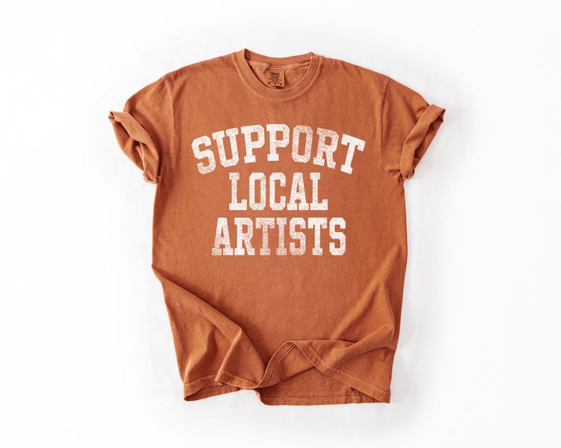 May include: A rust-colored t-shirt with the text "SUPPORT LOCAL ARTISTS" in a distressed white font. The shirt has rolled-up sleeves and a crew neck, perfect for casual wear. The shirt is made of a soft material.