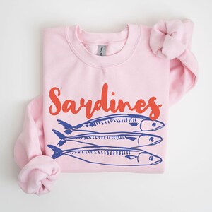 May include: Pink sweatshirt with the text "Sardines" in red and three blue sardines printed on the front.