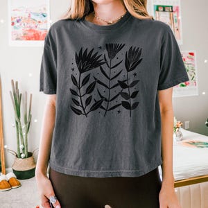 May include: A dark grey t-shirt with a black floral design, leaves and stars. The word "PEPPER" is printed in white at the bottom left. The shirt has a relaxed fit.