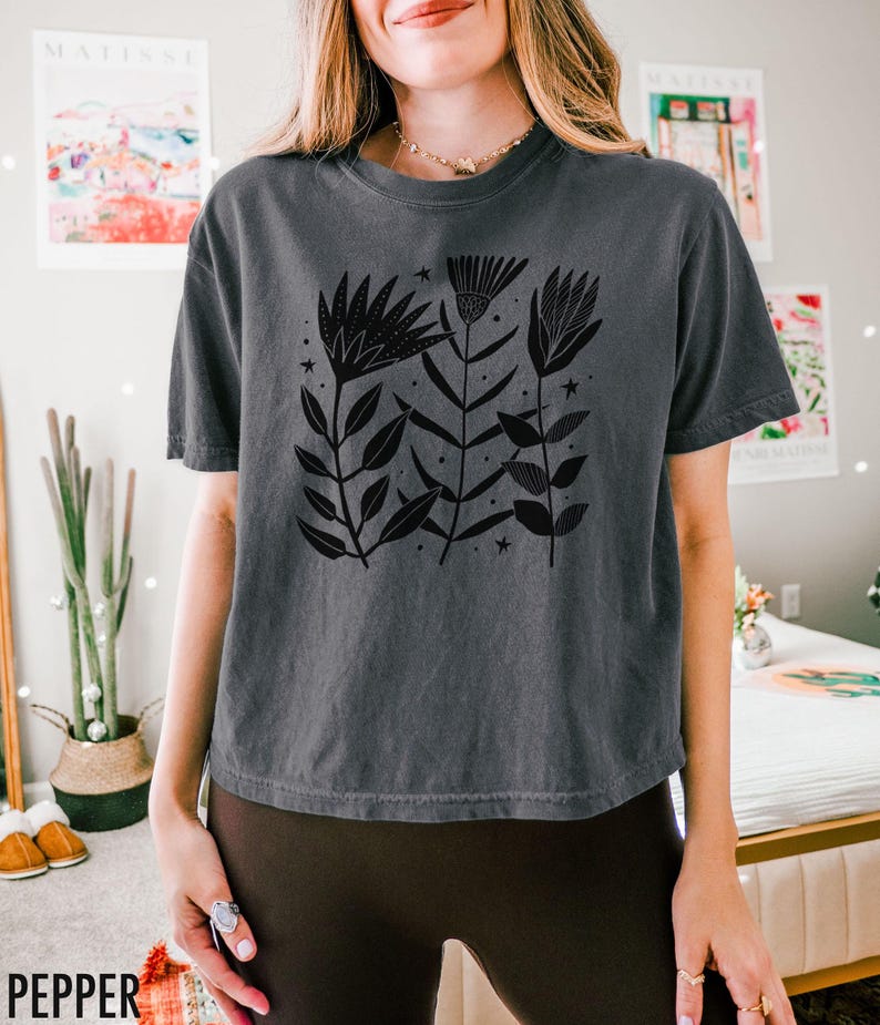 May include: A dark grey t-shirt with a black floral design, leaves and stars. The word "PEPPER" is printed in white at the bottom left. The shirt has a relaxed fit.