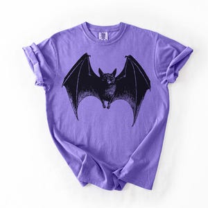 May include: A purple t-shirt featuring a black illustration of a bat with outstretched wings. The bat is centered on the shirt, with its wings spanning across the chest. The shirt has short sleeves and a crew neck.