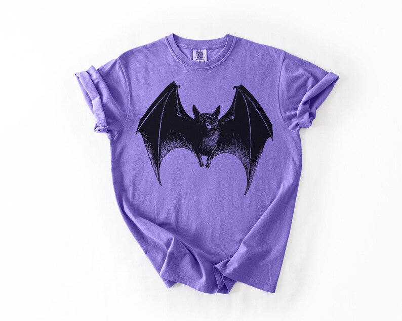 May include: A purple t-shirt featuring a black illustration of a bat with outstretched wings. The bat is centered on the shirt, with its wings spanning across the chest. The shirt has short sleeves and a crew neck.