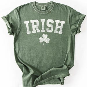 May include: Green t-shirt with the word "IRISH" in large, white, distressed letters and a white shamrock graphic. The shirt has rolled-up sleeves and a crew neck.