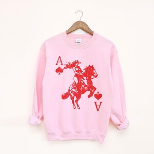 May include: A pale pink sweatshirt featuring a red graphic of a woman riding a horse, with the Ace of Spades and Ace of Hearts card symbols. The letter "A" is above the symbols. The sweatshirt is suspended from a wooden hanger.
