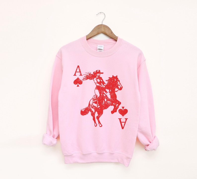 May include: A pale pink sweatshirt featuring a red graphic of a woman riding a horse, with the Ace of Spades and Ace of Hearts card symbols. The letter "A" is above the symbols. The sweatshirt is suspended from a wooden hanger.