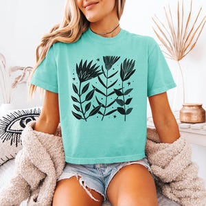 May include: A turquoise t-shirt with a black floral design. The graphic features stylized flowers, leaves, and stars. The short-sleeved shirt has a relaxed fit, suitable for casual wear.