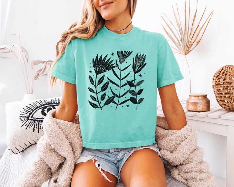 May include: A turquoise t-shirt with a black floral design. The graphic features stylized flowers, leaves, and stars. The short-sleeved shirt has a relaxed fit, suitable for casual wear.