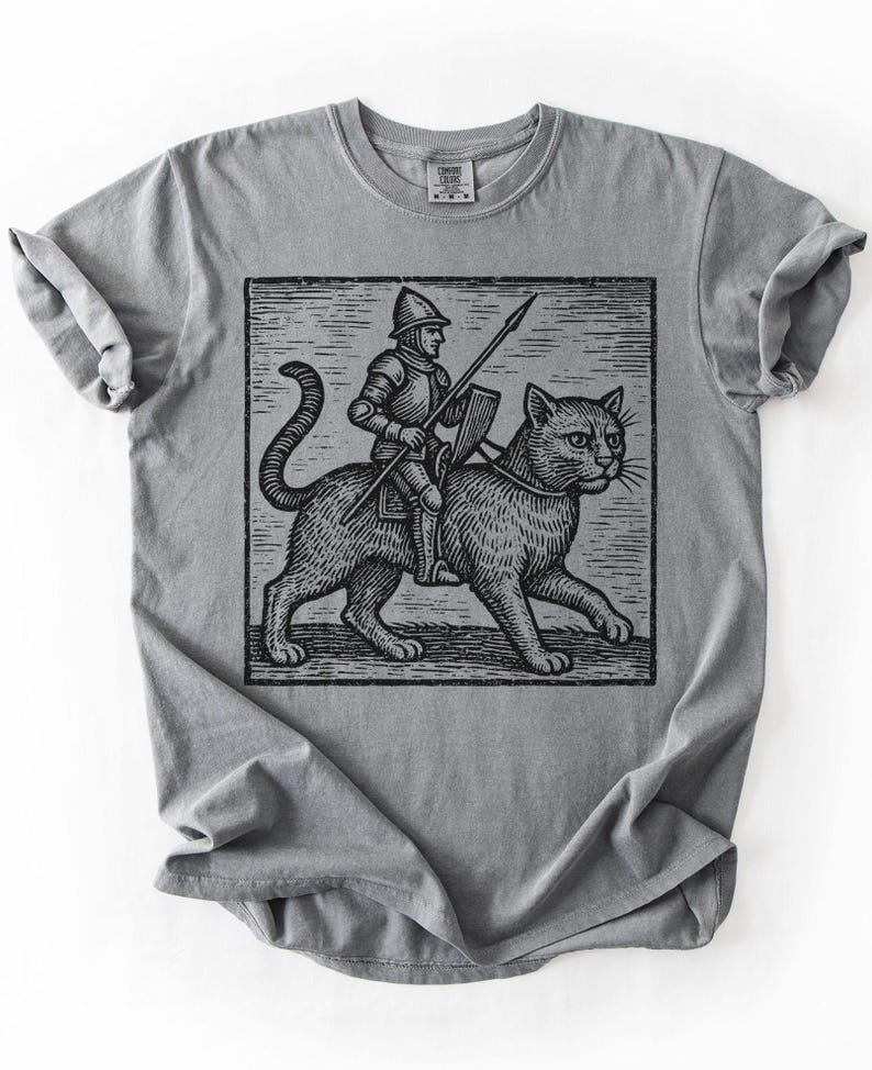 Puede incluir: Camiseta gris con una ilustraci&oacute;n en blanco y negro de un caballero montando un gato grande, sosteniendo una lanza y un escudo. El gr&aacute;fico est&aacute; enmarcado en un cuadrado. La camiseta es de material suave.