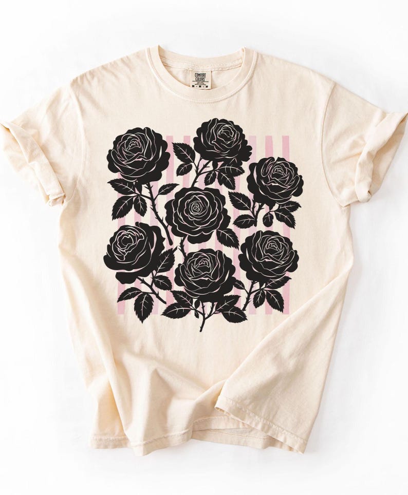 May include: Cream-colored t-shirt featuring a black rose graphic with pink vertical stripes. The graphic includes multiple detailed rose blooms and leaves. The shirt has a classic crew neck and rolled-up sleeves.