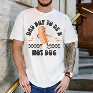 May include: White t-shirt with a retro graphic of a hot dog with the text "Bad Day To Be A Hot Dog".