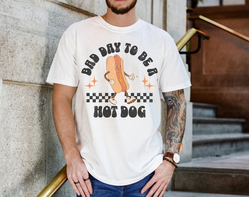 May include: White t-shirt with a retro graphic of a hot dog with the text "Bad Day To Be A Hot Dog".