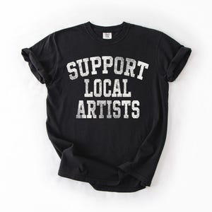 May include: Black t-shirt with the text "SUPPORT LOCAL ARTISTS" in distressed white lettering. The shirt has short sleeves and a crew neck. The shirt is made of a soft material.
