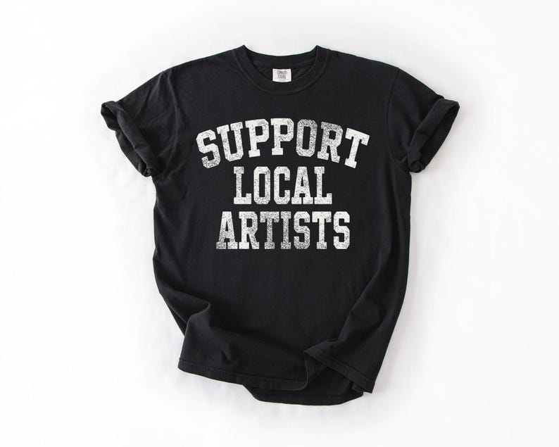 May include: Black t-shirt with the text "SUPPORT LOCAL ARTISTS" in distressed white lettering. The shirt has short sleeves and a crew neck. The shirt is made of a soft material.