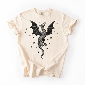 Op de afbeelding: Cr&egrave;mekleurig T-shirt met een gedetailleerd zwart drakenontwerp, uitgespreide vleugels en een lange staart. Kleine zwarte sterren omringen de draak. Het shirt heeft korte mouwen en een ronde hals.
