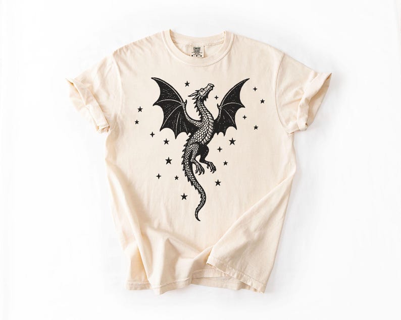 Op de afbeelding: Cr&egrave;mekleurig T-shirt met een gedetailleerd zwart drakenontwerp, uitgespreide vleugels en een lange staart. Kleine zwarte sterren omringen de draak. Het shirt heeft korte mouwen en een ronde hals.