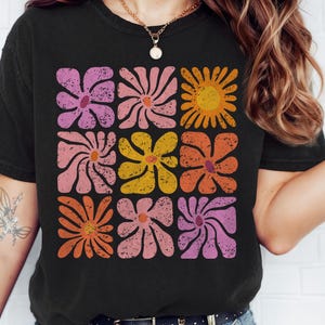 May include: A black t-shirt with a retro-style floral design in shades of pink, orange, and yellow. The flowers are arranged in a grid pattern and have a distressed, vintage look.
