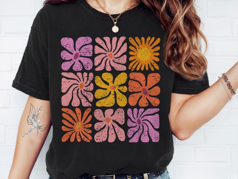 May include: A black t-shirt with a retro-style floral design in shades of pink, orange, and yellow. The flowers are arranged in a grid pattern and have a distressed, vintage look.