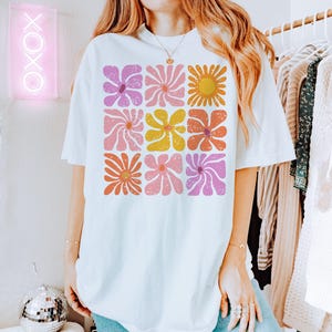 May include: White oversized t-shirt with a retro floral print featuring pink, yellow, orange, and purple flowers. The flowers are arranged in a grid pattern.