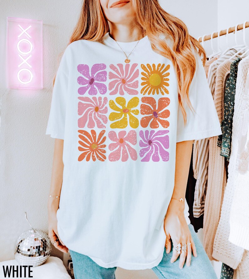 May include: White oversized t-shirt with a retro floral print featuring pink, yellow, orange, and purple flowers. The flowers are arranged in a grid pattern.
