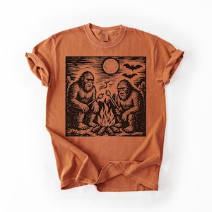 May include: Burnt orange t-shirt featuring a black graphic of two Bigfoot creatures roasting marshmallows over a campfire under a full moon. The design includes bats flying in the sky. The shirt has a crew neck and short sleeves.