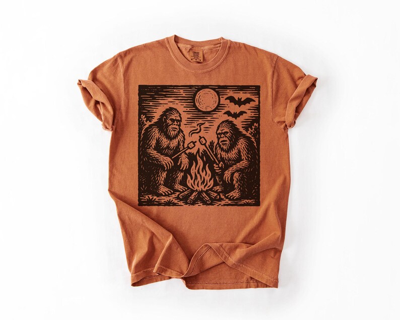 May include: Burnt orange t-shirt featuring a black graphic of two Bigfoot creatures roasting marshmallows over a campfire under a full moon. The design includes bats flying in the sky. The shirt has a crew neck and short sleeves.