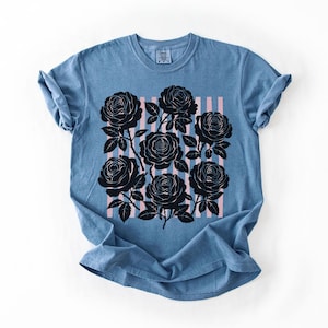 May include: A blue t-shirt featuring a black rose graphic with pink vertical stripes. The roses are detailed with stems and leaves. The shirt has rolled-up sleeves and a crew neck, and is made of a soft material.