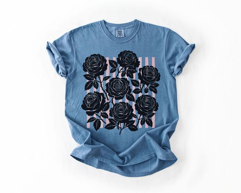 May include: A blue t-shirt featuring a black rose graphic with pink vertical stripes. The roses are detailed with stems and leaves. The shirt has rolled-up sleeves and a crew neck, and is made of a soft material.