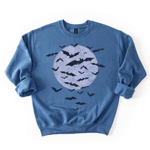 May include: A blue crew neck sweatshirt with a distressed graphic of a full moon and bats. The bats are black, and the moon is a lighter blue. The sweatshirt has rolled cuffs. The design is Halloween themed.