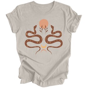 Puede incluir: Una camiseta de color polvo de brezo con un dise&ntilde;o gr&aacute;fico de dos serpientes entrelazadas alrededor de un s&iacute;mbolo de sol y luna. Las serpientes son marrones con detalles dorados.