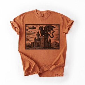 May include: Burnt orange t-shirt featuring a black graphic of a cat-like creature with wings atop a castle, with a UFO in the sky. The design is a woodcut style illustration.