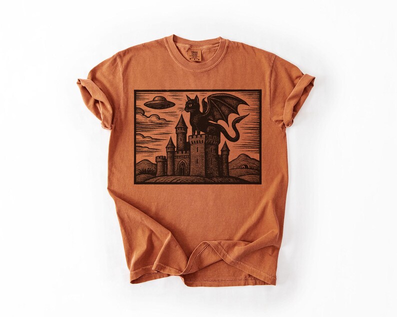 May include: Burnt orange t-shirt featuring a black graphic of a cat-like creature with wings atop a castle, with a UFO in the sky. The design is a woodcut style illustration.