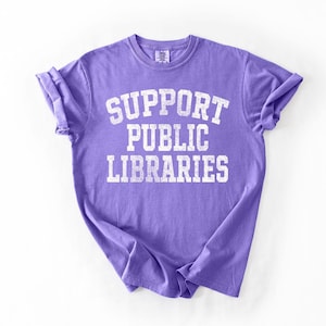 May include: A purple t-shirt with the text "SUPPORT PUBLIC LIBRARIES" in white, block letters. The shirt has rolled-up sleeves and a crew neck. The fabric has a slightly faded appearance.