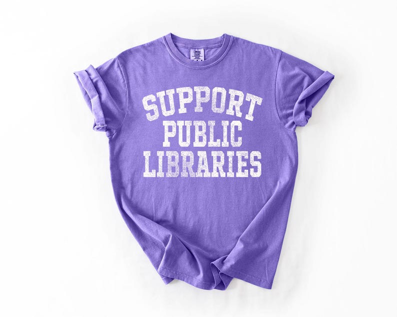 May include: A purple t-shirt with the text "SUPPORT PUBLIC LIBRARIES" in white, block letters. The shirt has rolled-up sleeves and a crew neck. The fabric has a slightly faded appearance.