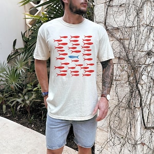 May include: A cream-colored t-shirt with a graphic of red fish and one blue fish. The fish are arranged in rows.