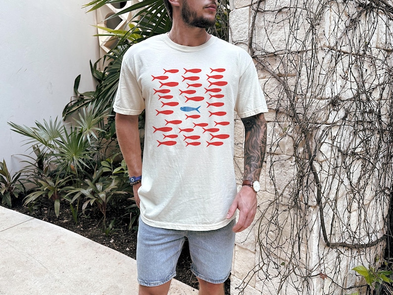 May include: A cream-colored t-shirt with a graphic of red fish and one blue fish. The fish are arranged in rows.