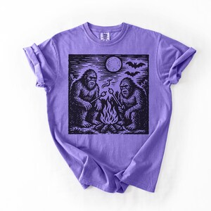May include: A purple t-shirt featuring a black and white graphic of two Bigfoot creatures roasting marshmallows over a campfire under a full moon. Bats are flying in the sky above the scene.