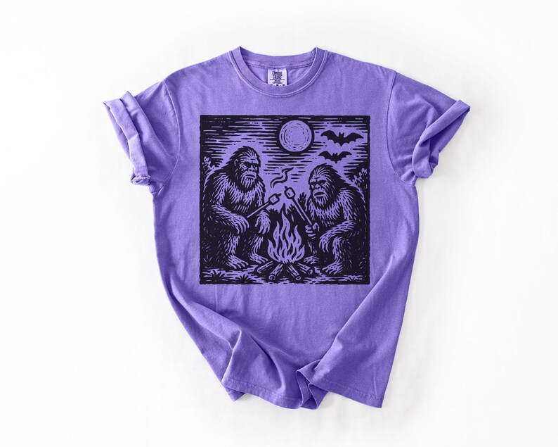 May include: A purple t-shirt featuring a black and white graphic of two Bigfoot creatures roasting marshmallows over a campfire under a full moon. Bats are flying in the sky above the scene.