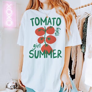 May include: White t-shirt with a graphic of red tomatoes on a vine with green leaves. The text "TOMATO girl SUMMER" is printed on the shirt. The text "Italy" is printed on the right side of the tomato graphic.