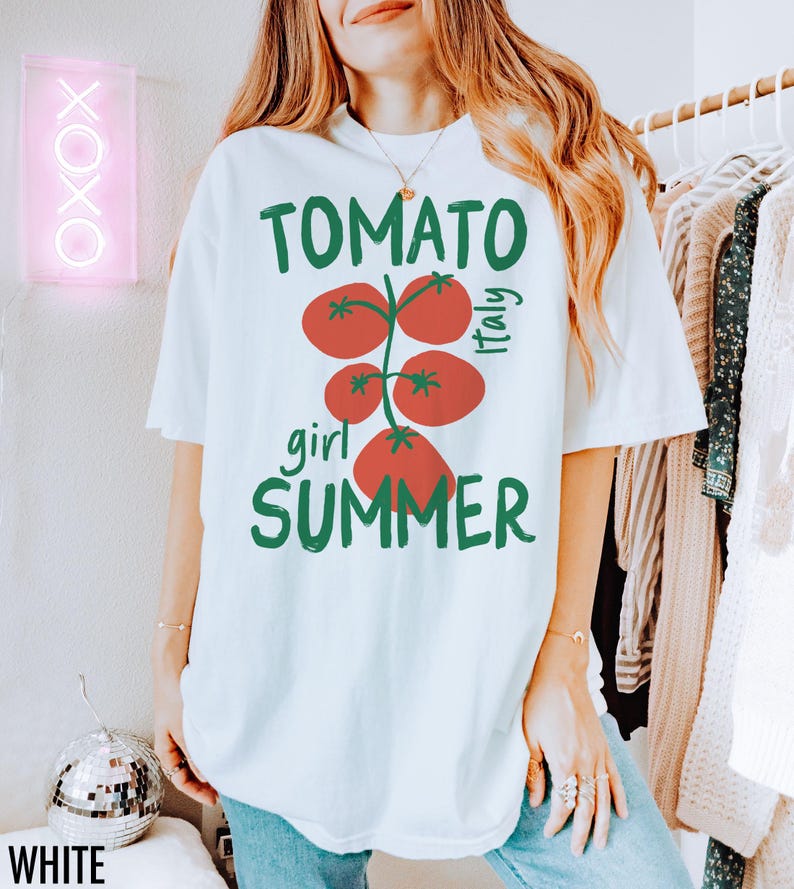 May include: White t-shirt with a graphic of red tomatoes on a vine with green leaves. The text "TOMATO girl SUMMER" is printed on the shirt. The text "Italy" is printed on the right side of the tomato graphic.
