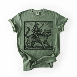 Puede incluir: Camiseta verde oliva con una ilustraci&oacute;n en blanco y negro de un caballero montado en un gato, sosteniendo una lanza y un escudo. El gr&aacute;fico est&aacute; enmarcado en un rect&aacute;ngulo negro. La camiseta tiene cuello redondo y mangas enrolladas.