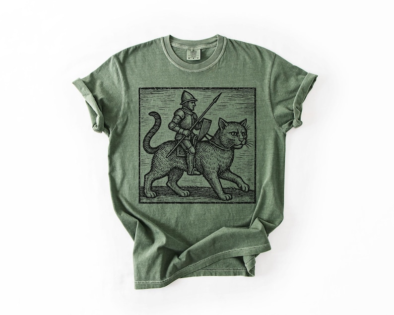 Puede incluir: Camiseta verde oliva con una ilustraci&oacute;n en blanco y negro de un caballero montado en un gato, sosteniendo una lanza y un escudo. El gr&aacute;fico est&aacute; enmarcado en un rect&aacute;ngulo negro. La camiseta tiene cuello redondo y mangas enrolladas.